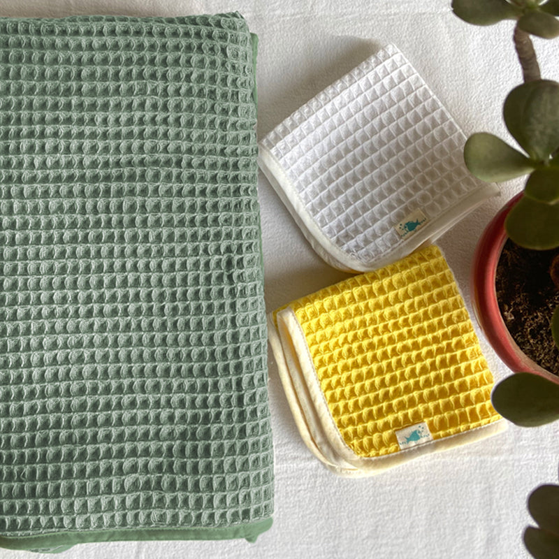 Three waffle-textured towels in green, white, and yellow on a light surface with a plant in the corner.