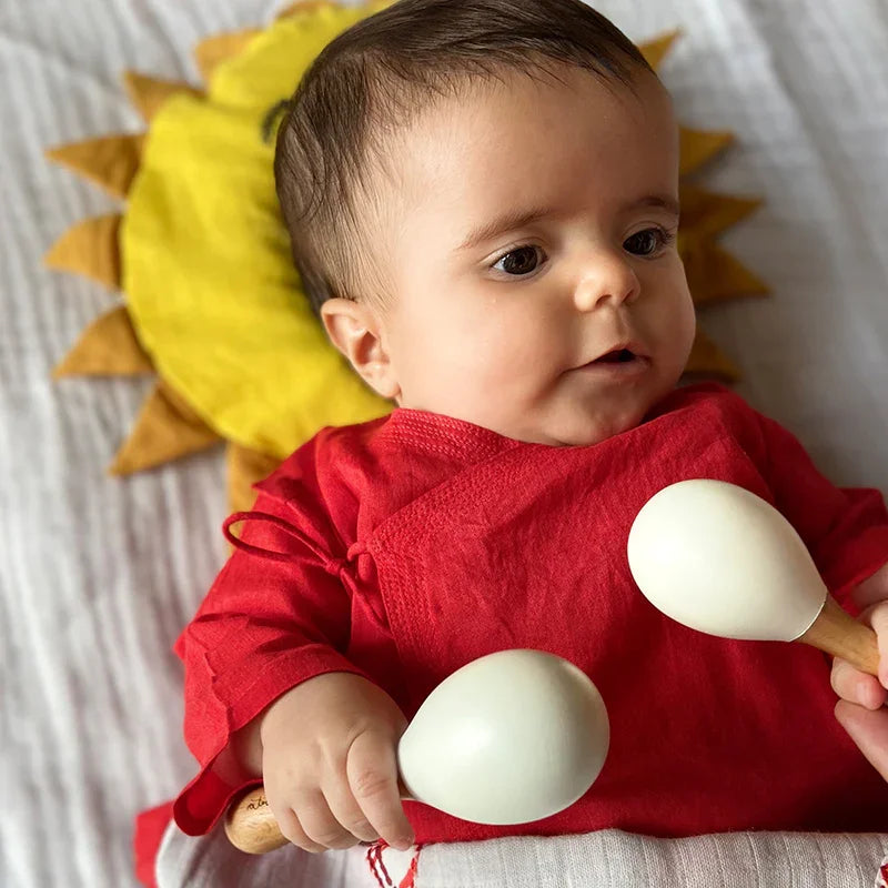 Baby in a red outfit holding wooden rattle toys with a sun-shaped pillow in the background.