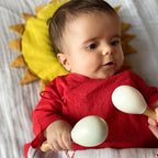 Baby in a red outfit holding wooden rattle toys with a sun-shaped pillow in the background.